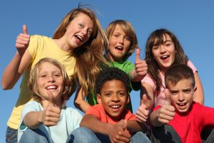 A group of six diverse children all smiling, huddled together with their thumbs up.