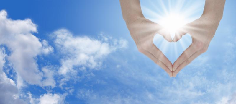 hands held in heart shape in front of sun and blue and cloudy sky